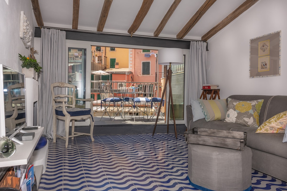 The living area features patterned tiles on the floor, a comfortable gray sofa, and a decorative light fixture on a wooden stand. Curtains frame a large glass door that opens to a terrace with outdoor seating, offering views of the surrounding colorful buildings.