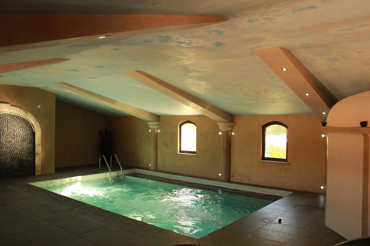 An indoor pool area is shown with a tranquil turquoise water surface. Soft light illuminates the space, highlighting the textured wall finishes and subtle architectural details. Arched windows provide views of the exterior, contributing to a serene ambiance.