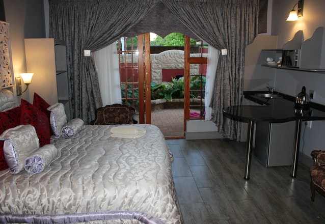 A spacious guest room features a large, elegantly patterned bed with decorative pillows. Large windows offer views of the garden, framed by soft, flowing drapes. A compact kitchen area and a small seating arrangement increase functionality, while the flooring has a natural wood look.