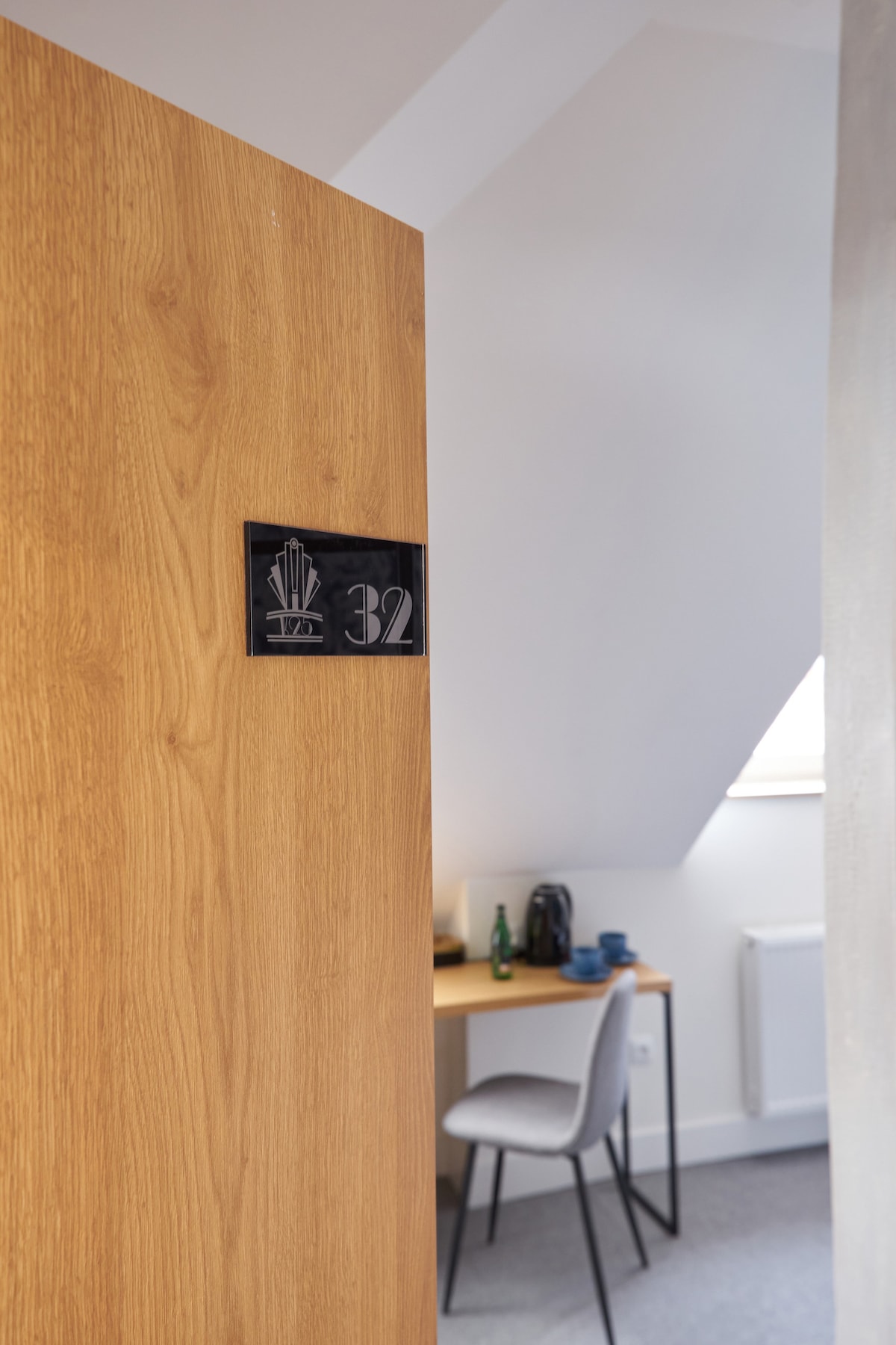 A wooden door is presented at an angle, showcasing the room number 32. Beyond the door, a comfortable workspace is visible, featuring a chair and a desk equipped with a kettle and glass bottles. Natural light enters through a window, illuminating the inviting space.