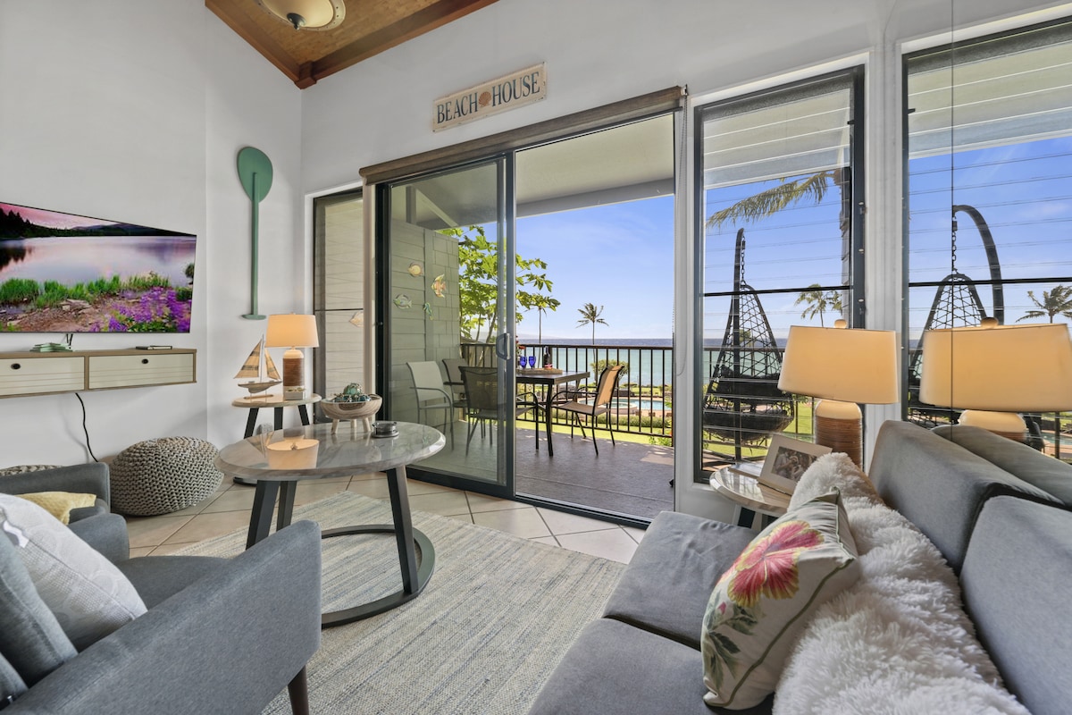 A bright living area features a comfortable sofa and modern decor, with large sliding glass doors leading to a private lanai. A TV is mounted on the wall, and the ocean view is complemented by outdoor seating surrounded by palm trees.