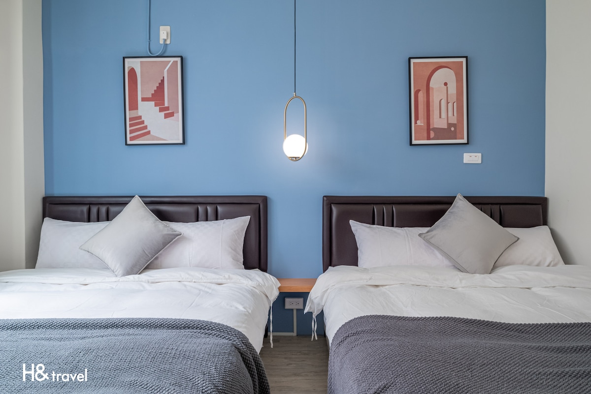 A well-organized sleeping area features two spacious double beds with neutral bed linens. Behind the beds, a calming blue wall is adorned with framed artwork. A modern pendant light hangs overhead, contributing to the room's functional design.