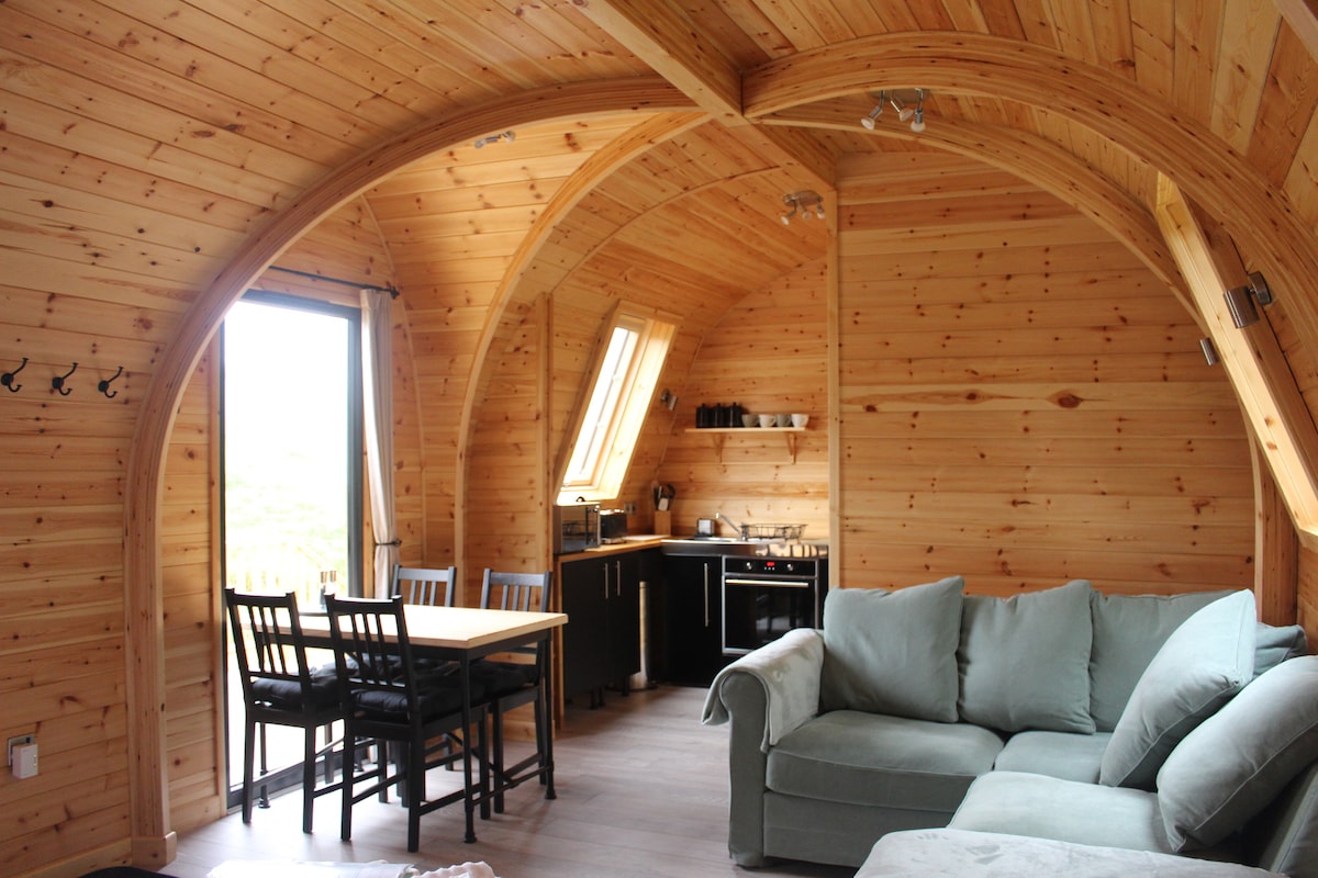 The interior of the pod features a vaulted wooden ceiling, enhancing the spacious feel. A well-furnished kitchen is visible, alongside a dining table with chairs. An L-shaped couch offers a cozy seating area. Large windows allow natural light and scenic views.