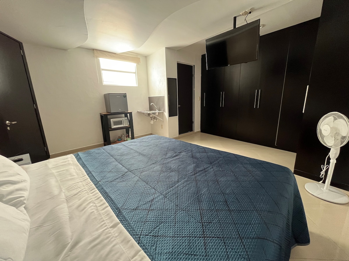 A spacious sleeping area features a large bed with a textured blue cover. Dark wooden cabinets provide ample storage, while a mounted television hangs on the wall. Natural light enters through a small window, and a small refrigerator is placed on a desk next to a wall fan.