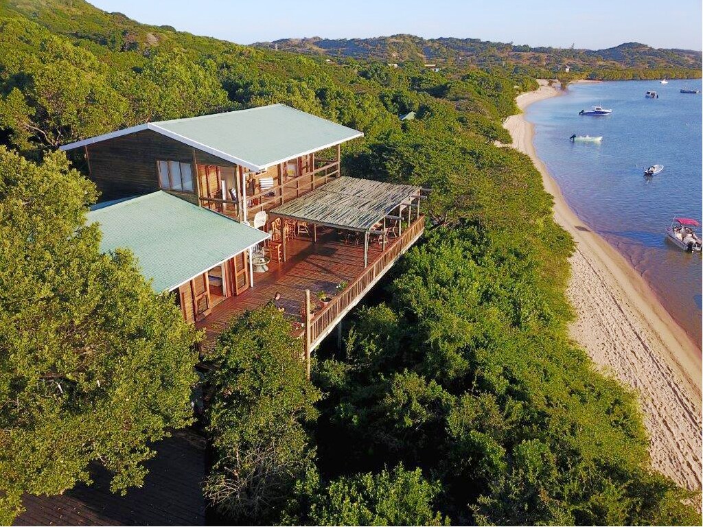 The house is nestled among lush greenery, featuring a spacious deck that extends into the surroundings. The two-story structure showcases a large roof and expansive windows, providing views of the calm waters and sandy beach nearby, with boats anchored in the distance.