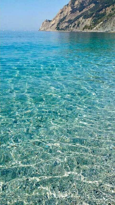 The image captures crystal-clear water gently lapping at a pebbled shoreline. Sunlight reflects off the surface, creating a shimmering effect. In the background, a rocky coastal line rises, partially covered by greenery, stretching towards a clear blue sky.