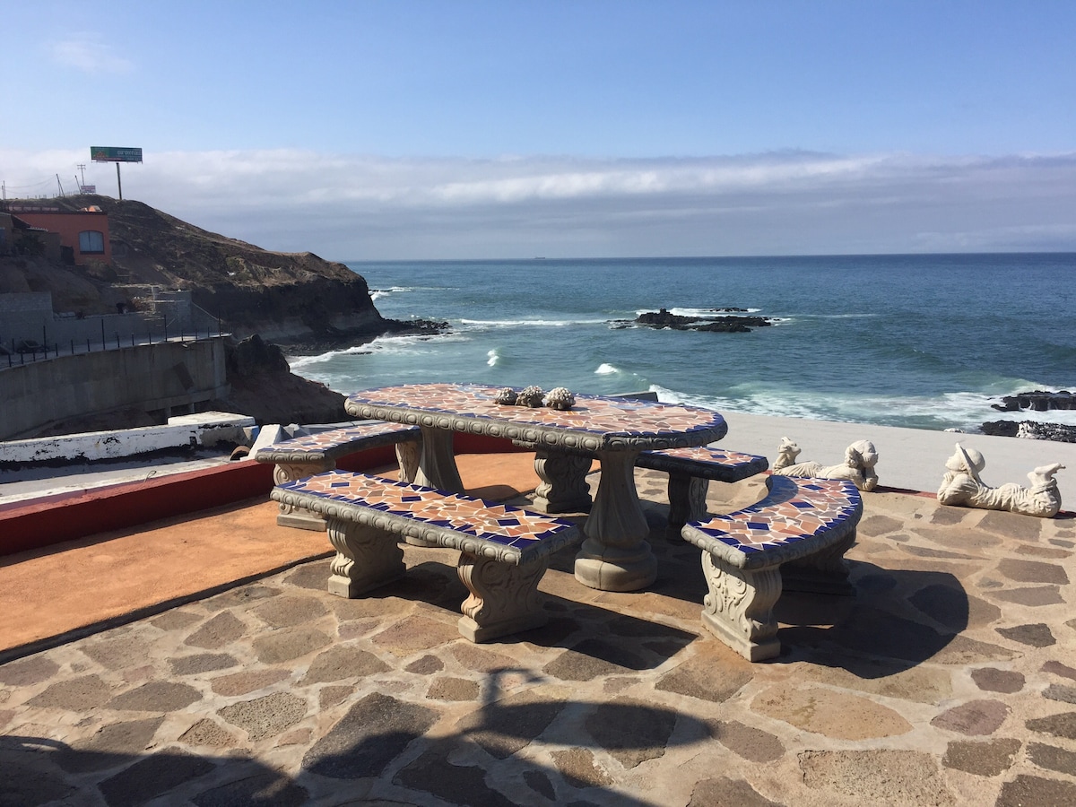 A large outdoor dining area features a mosaic-tiled table supported by stone bases, surrounded by benches on a stone patio. The ocean waves crash against the nearby rocks, and coastal scenery extends along the horizon under a clear blue sky.