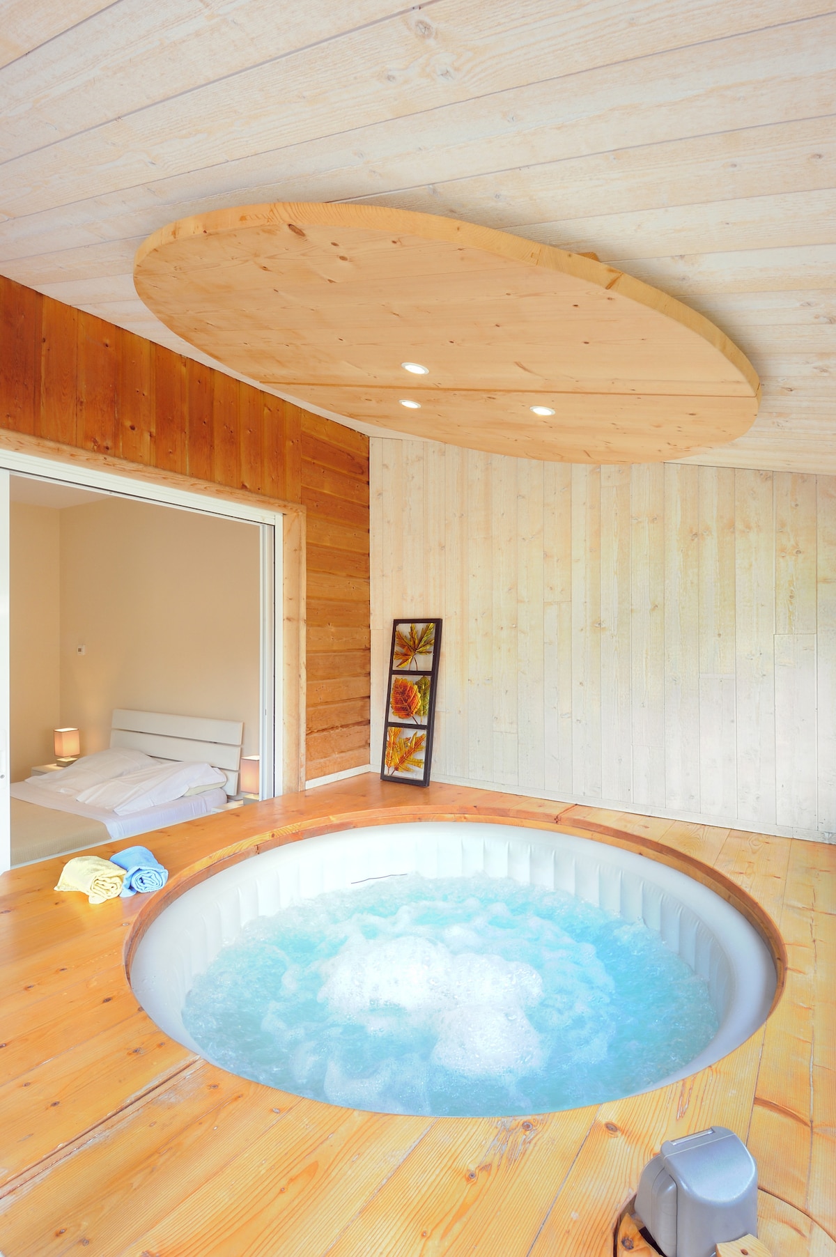 A circular spa is positioned in a wooden-floored room, featuring gentle bubbling water. The light wood walls are complemented by a round wooden ceiling with recessed lighting. A doorway leads to a softly lit sleeping area, with towels placed neatly on the spa's edge.