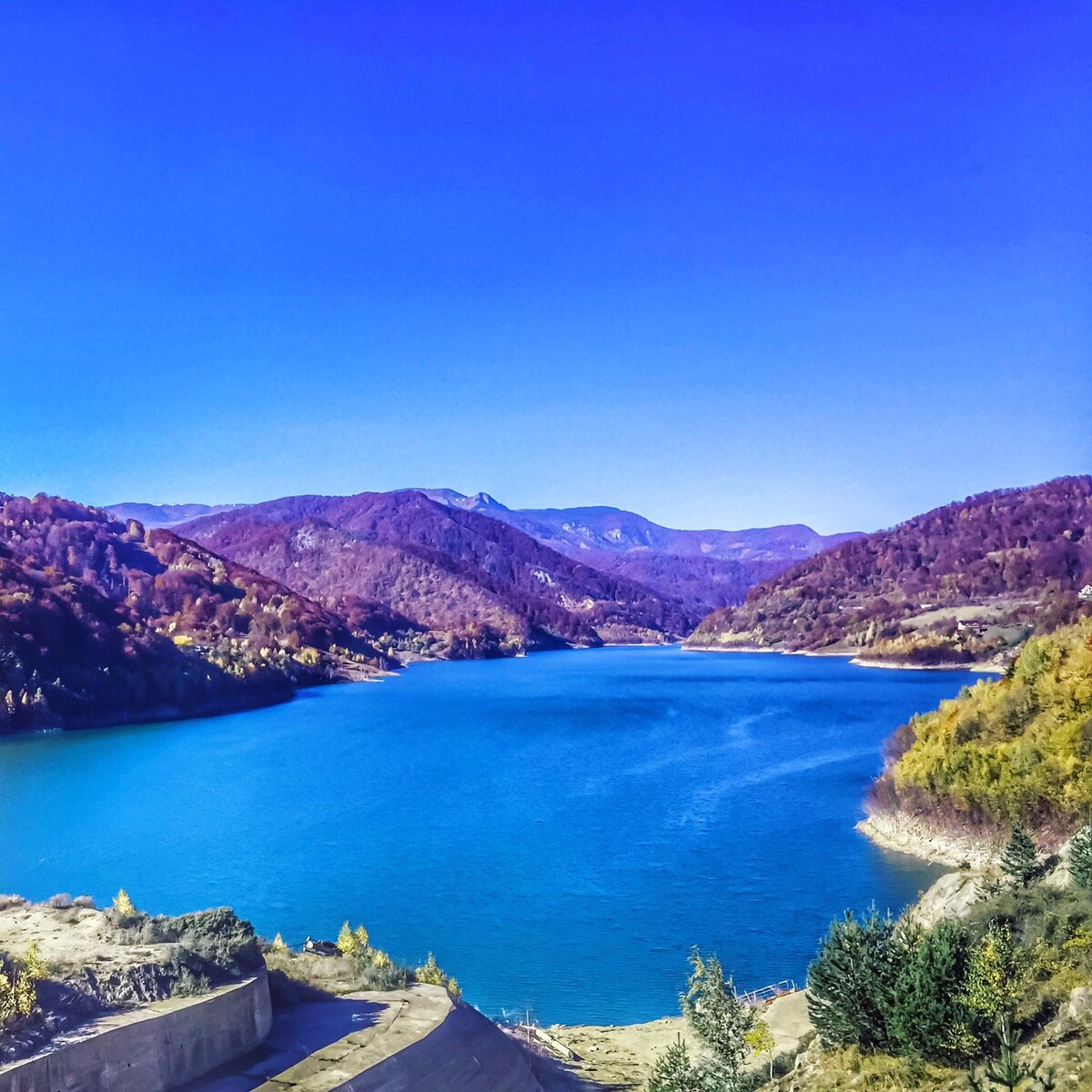 An expansive view of a tranquil lake is captured, surrounded by rolling hills and autumn foliage. The water reflects a range of blue shades under a clear sky, with mountains visible in the background, creating a serene natural landscape.