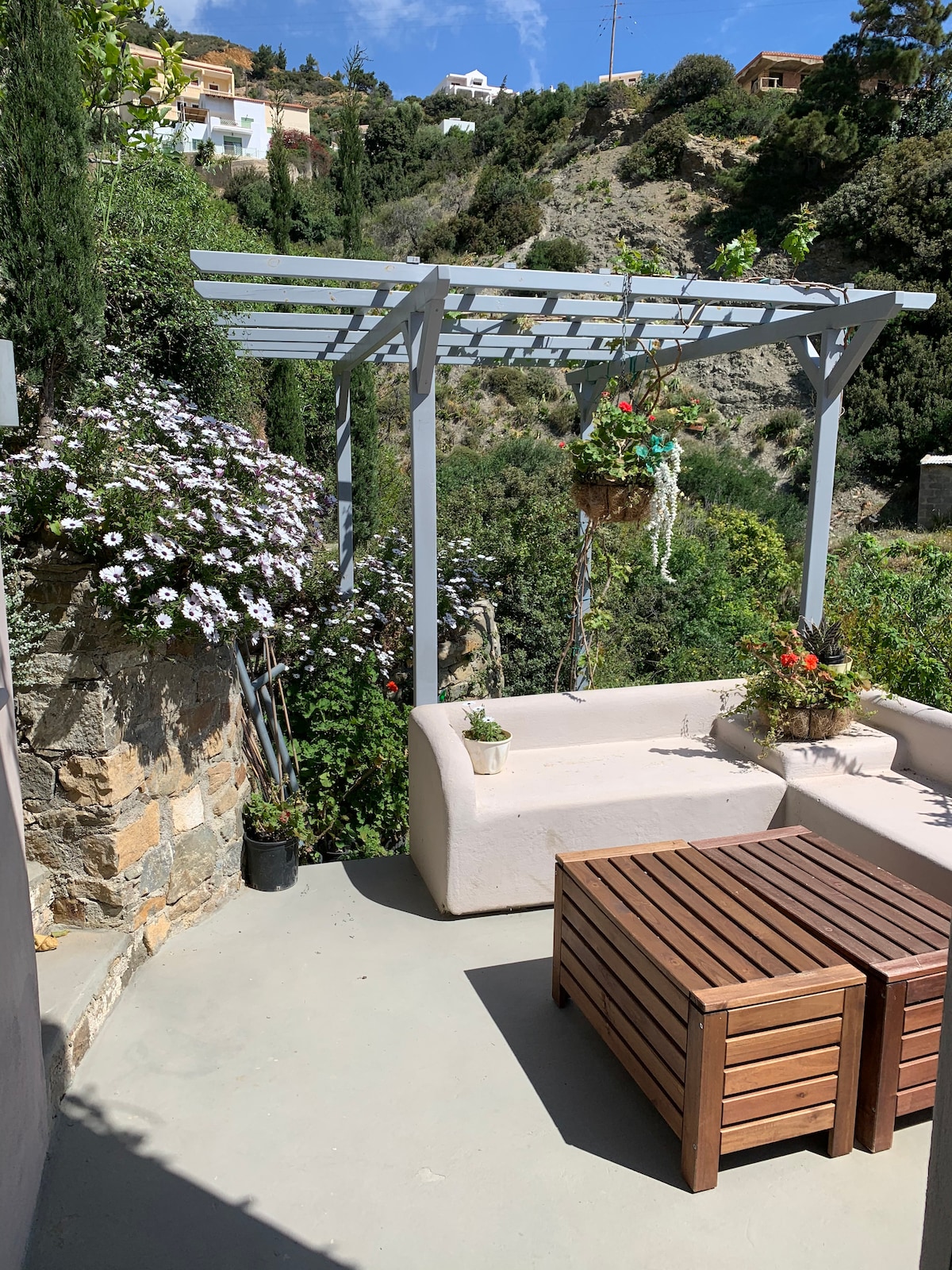 A shaded seating area is nestled under a wooden pergola, surrounded by blooming flowers and lush greenery. A compact wooden table sits in front of a low stone wall, providing a peaceful space for relaxation or dining in the outdoors.