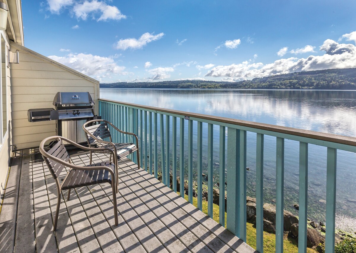 A private balcony offers views of the water and surrounding landscape. Two chairs are positioned for relaxation, accompanied by a gas barbecue. The sunlight casts a patterned shadow across the wooden deck, enhancing the serene atmosphere.