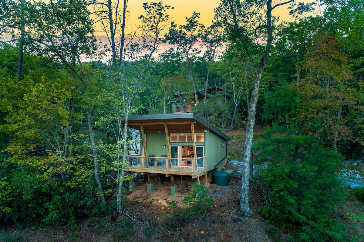 The modern cabin is nestled among trees, elevated on stilts, and features a spacious covered deck that invites outdoor enjoyment. Large windows reflect the greenery, allowing for natural light to enhance the welcoming design. A serene forest backdrop complements the structure.
