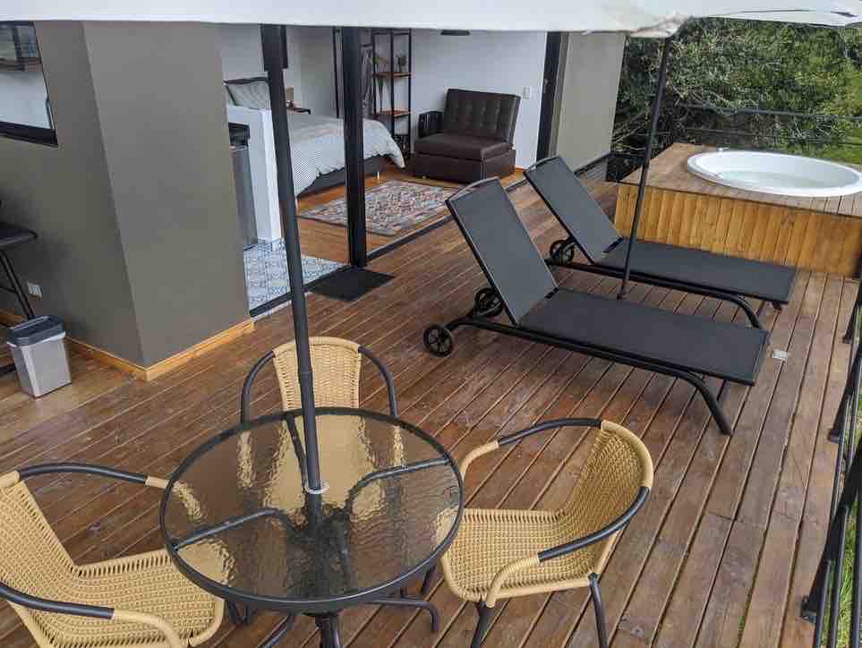 An elevated terrace is featured with a round table surrounded by four wicker chairs. Two lounge chairs are positioned for sunbathing, while a wooden structure houses a compact hot tub nearby. Access to the interior space and a cozy sleeping area is visible through glass doors.
