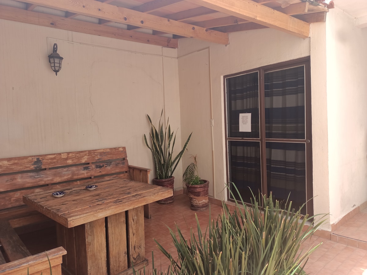 A shaded patio area features a rustic wooden table and bench, complemented by potted plants. Light filters through the overhang, creating a gentle ambiance. The space is bordered by textured walls and glass doors, enhancing the connection to the indoors.