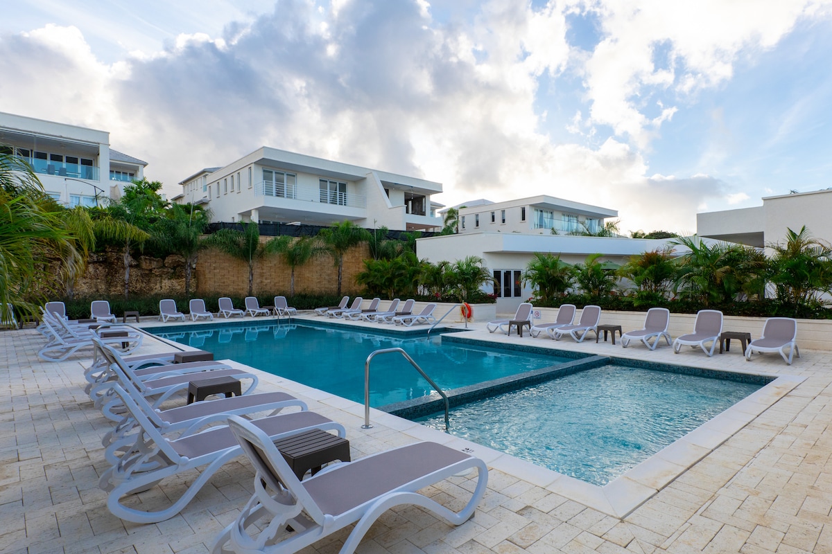 A communal swimming pool area features a large rectangular pool surrounded by multiple lounge chairs. Palm trees and greenery offer natural accents around the space, while modern white buildings are visible in the background. The sky is partly cloudy, contributing to a serene atmosphere.