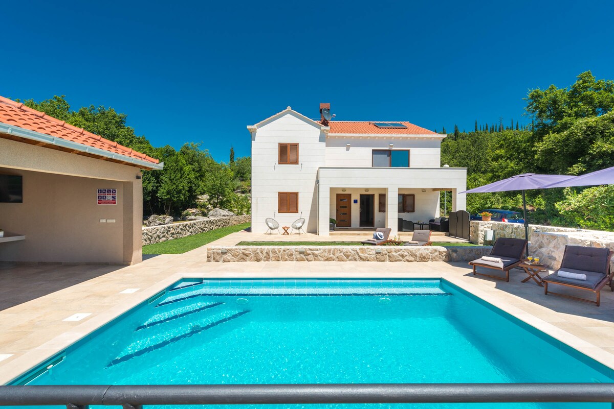 A private swimming pool is framed by a stone terrace, reflecting the bright blue sky. The multi-story villa, featuring a mix of light-colored walls and a red tiled roof, stands in the background among lush greenery. Seating areas are arranged around the pool for relaxation.