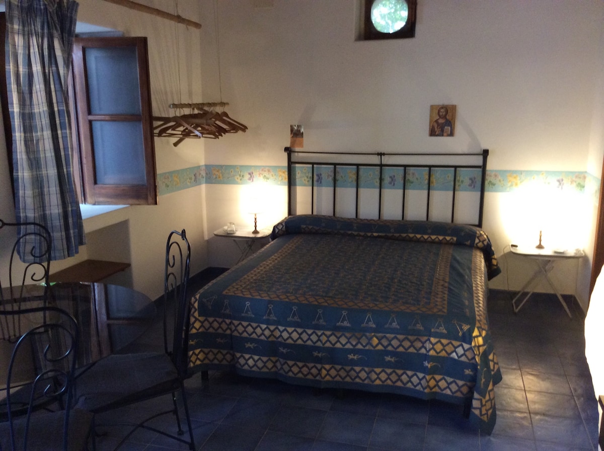 A spacious room features a double bed with a patterned blanket. Two bedside tables with lamps are placed on either side. A small table and chairs are situated near a window, with wooden hangers for clothing visible in the background.
