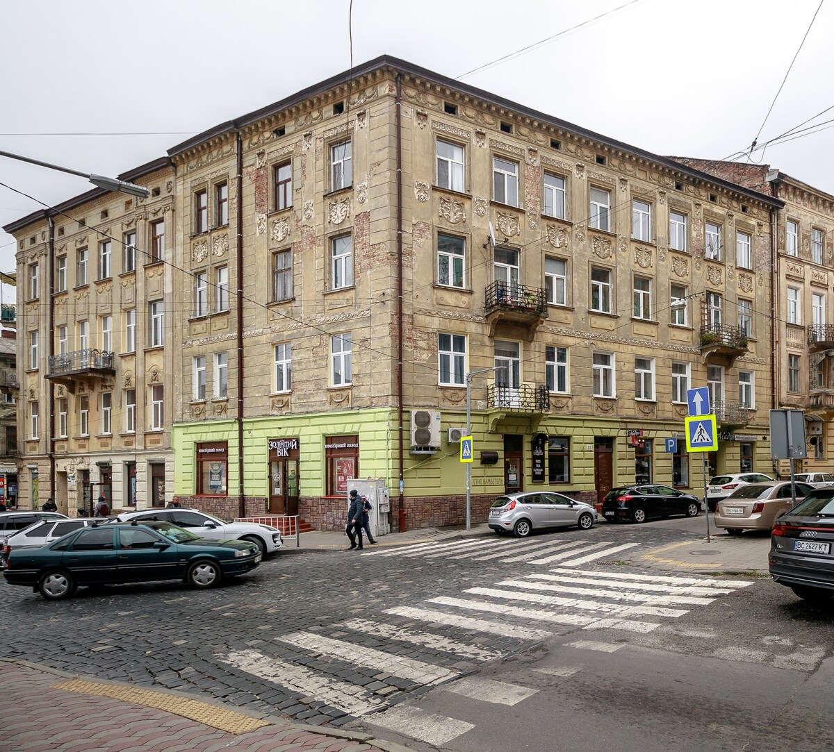 A historic building with intricate façade details is showcased, featuring multiple stories and balconies. The scene captures an intersection with vehicles parked along the street and pedestrians crossing at a designated crosswalk. A tranquil urban atmosphere is evident from the cobblestone pavement.