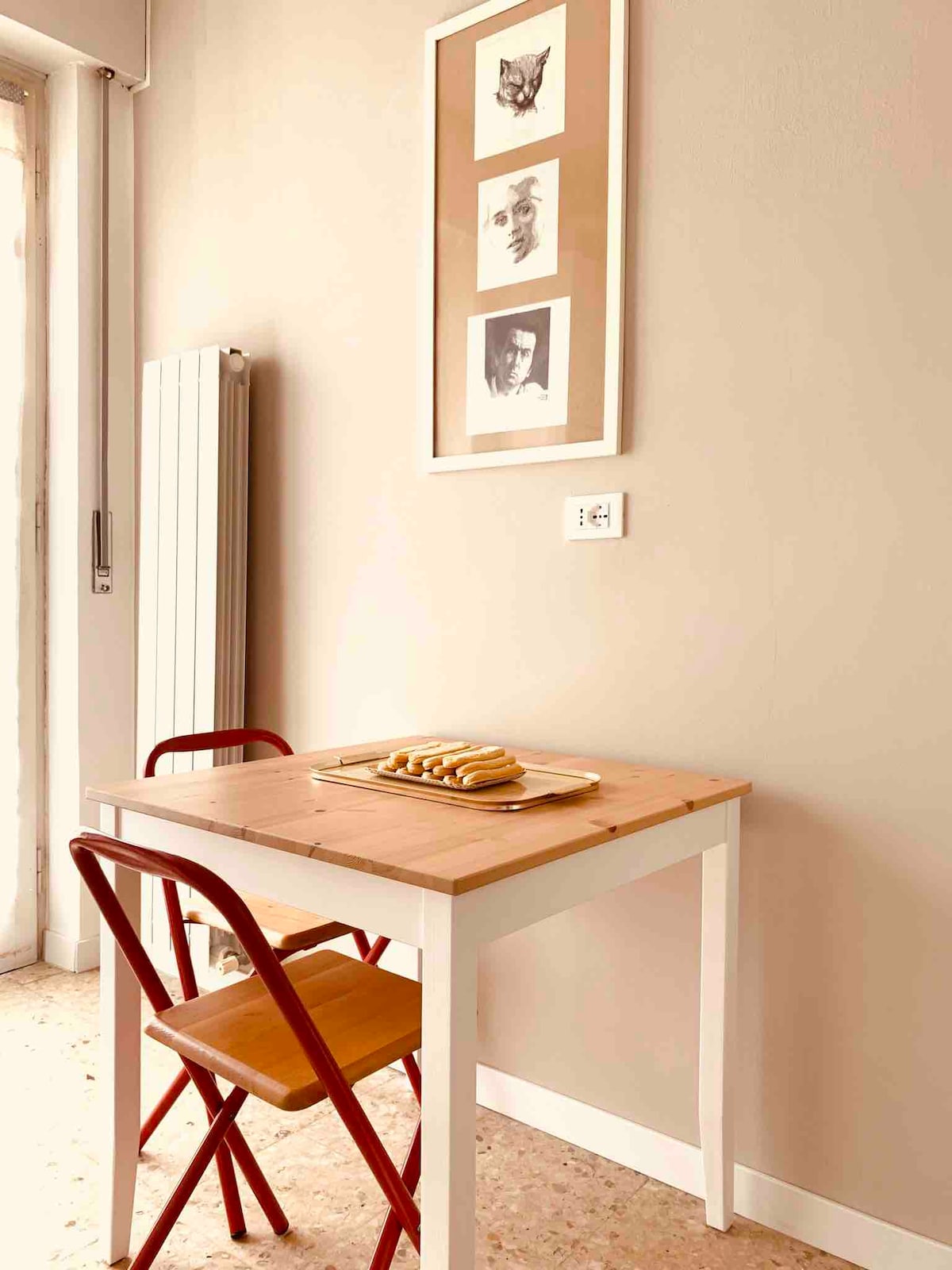 A small dining area is depicted with a light wooden table and two chairs, one red and one beige. A simple plate of pastries rests on the table. The wall features a decorative framed art piece, adding a touch of visual interest to the neutral background.