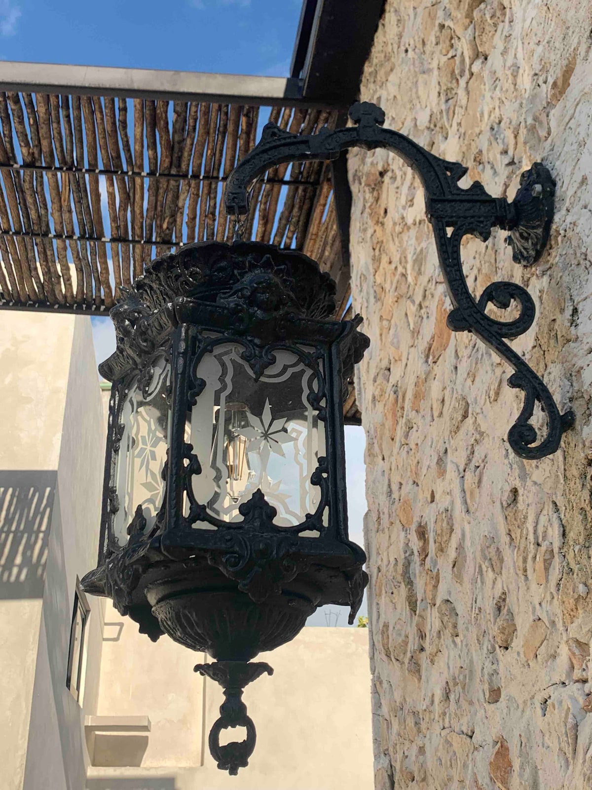 An ornate black lantern is mounted on a textured stone wall. The lantern features intricate detailing and glass panels, allowing light to shine through. Above, a thatched roof provides shade, creating a contrast between the natural materials and the decorative fixture.