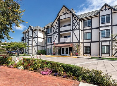 The exterior of the resort showcases a charming three-story building with a traditional design featuring timber accents. A well-kept landscape with colorful flower beds and neatly arranged pathways enhances the inviting entrance. Clear skies are visible above, contributing to a bright and welcoming atmosphere.