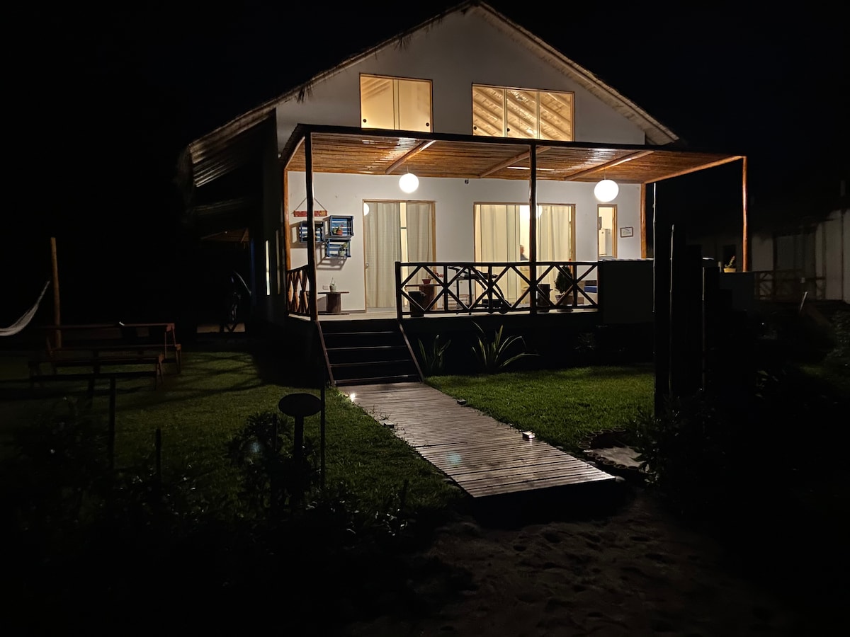 The exterior of a cabin is illuminated at night, showcasing a spacious deck with several hanging lights. Large windows reveal a warm glow from the interior. A wooden pathway leads to the entrance, surrounded by well-maintained greenery and a hammock nearby.