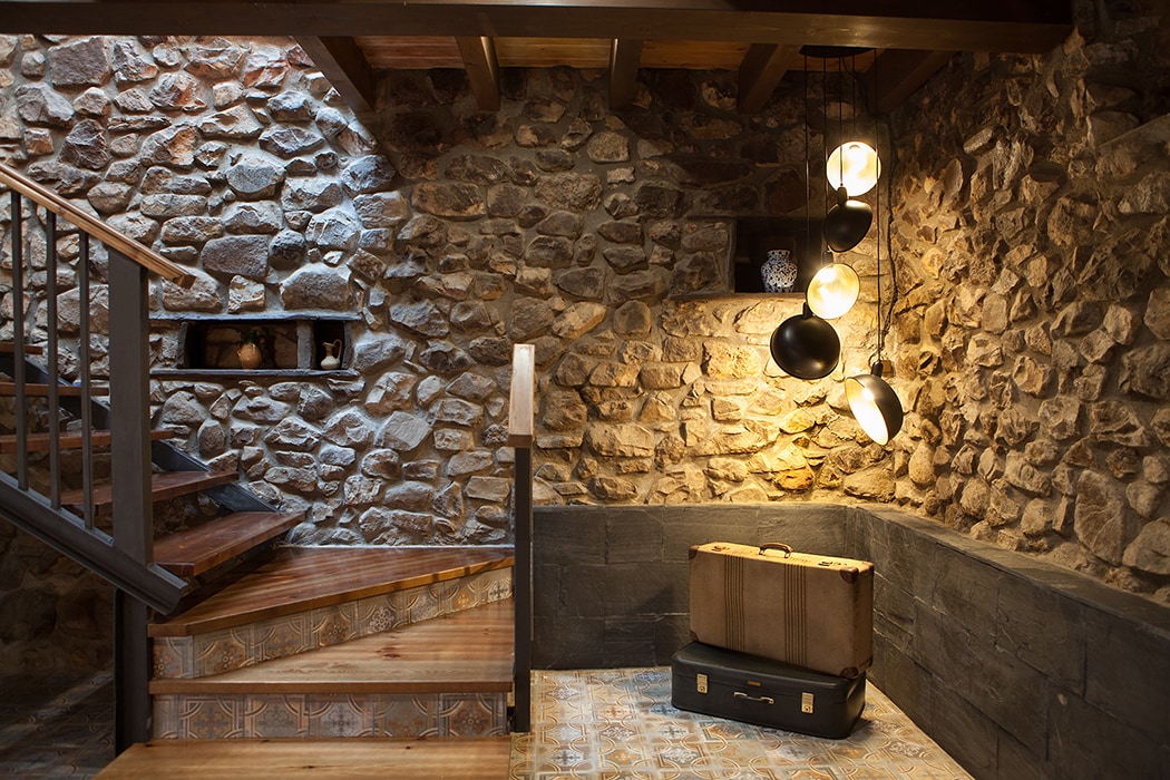 A stairway with wooden steps ascends alongside a textured stone wall. Soft lighting is provided by a cluster of hanging lamps. A vintage suitcase rests on a patterned tile floor, with a shelf above showcasing decorative items.