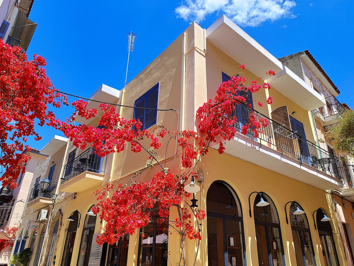 A corner of a charming building is highlighted by vibrant red bougainvillea cascading over the balcony. The bright yellow facade contrasts with the clear blue sky, creating an inviting detail of the picturesque neighborhood.