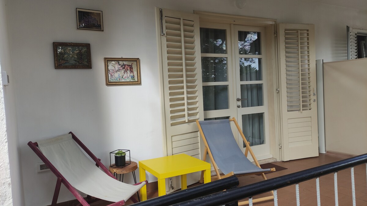 A covered terrace area features two reclining chairs positioned beside a small yellow table. The French doors are framed by white shutters, allowing light to enter the space. A potted plant sits on the table, enhancing the outdoor relaxation setting.