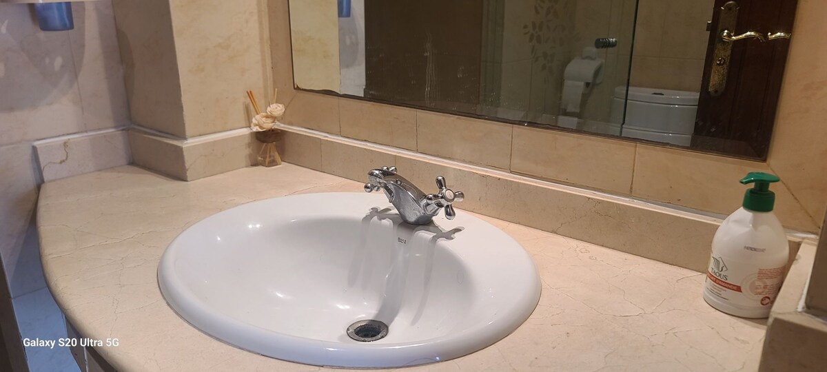 A clean and well-maintained bathroom sink is shown, featuring a polished white basin and a chrome faucet. A soap dispenser is positioned nearby, and a large mirror reflects the sink area. The countertop is finished in light marble, contributing to a tidy appearance.