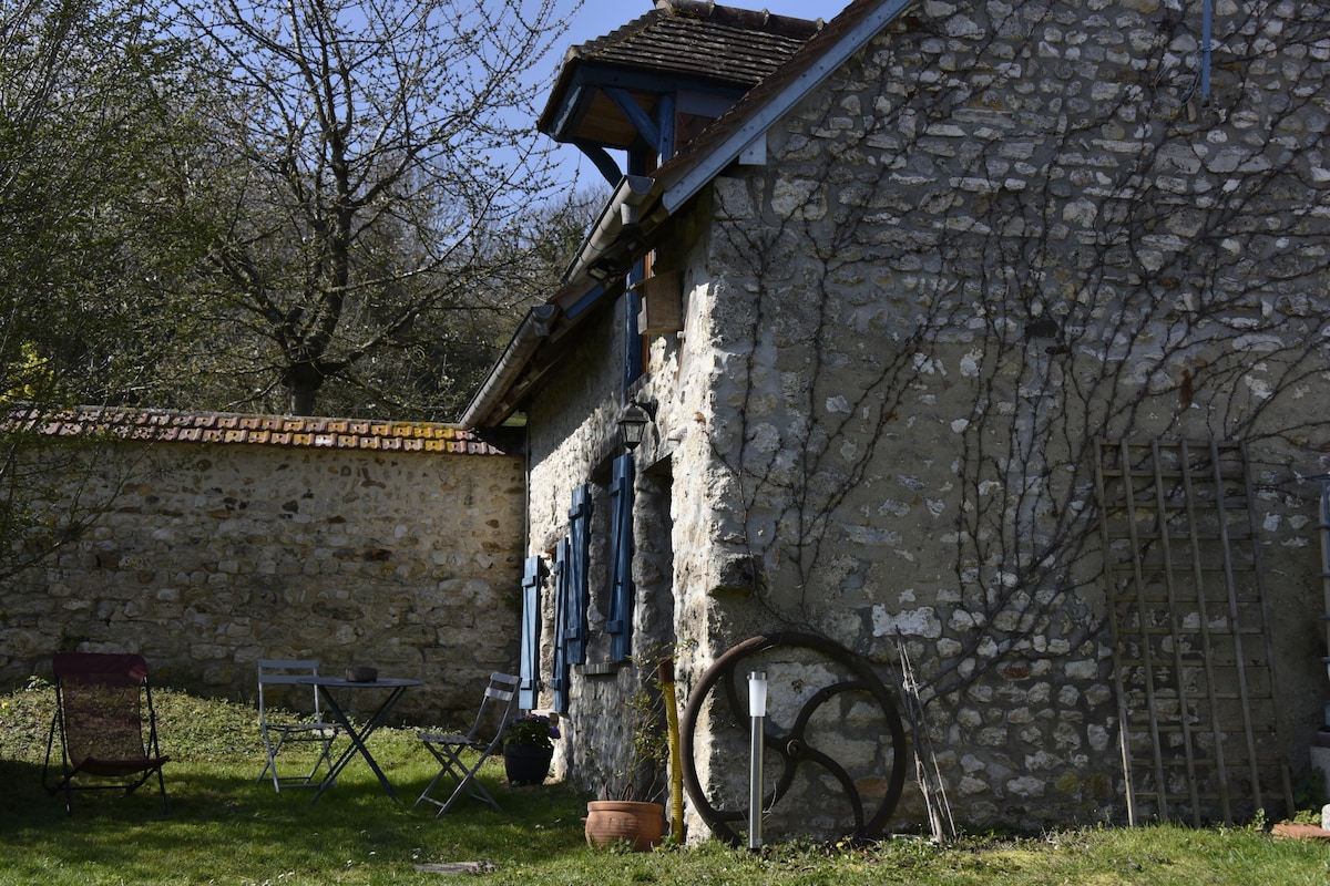 A charming stone cottage is seen surrounded by greenery, with blue-shuttered windows and decorative wooden elements. A small outdoor seating area with two chairs and a table is positioned on the grass, inviting relaxation in a peaceful setting.