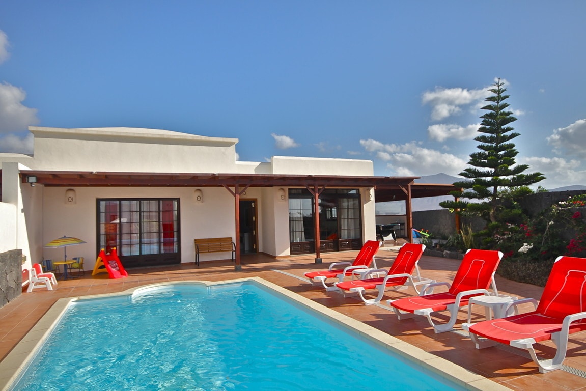 An outdoor area features a bright blue pool surrounded by red lounge chairs. A covered terrace extends from the house, with large windows providing visibility into the interior. Lush greenery and colorful plants frame the space, creating an inviting environment.