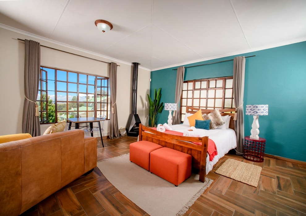 An open-plan studio apartment features a comfortable bed with colorful decorative pillows. Two lamps illuminate the room, which includes a cozy seating area with a leather couch and an orange bench. Large windows allow natural light to fill the space, complemented by warm wooden flooring.
