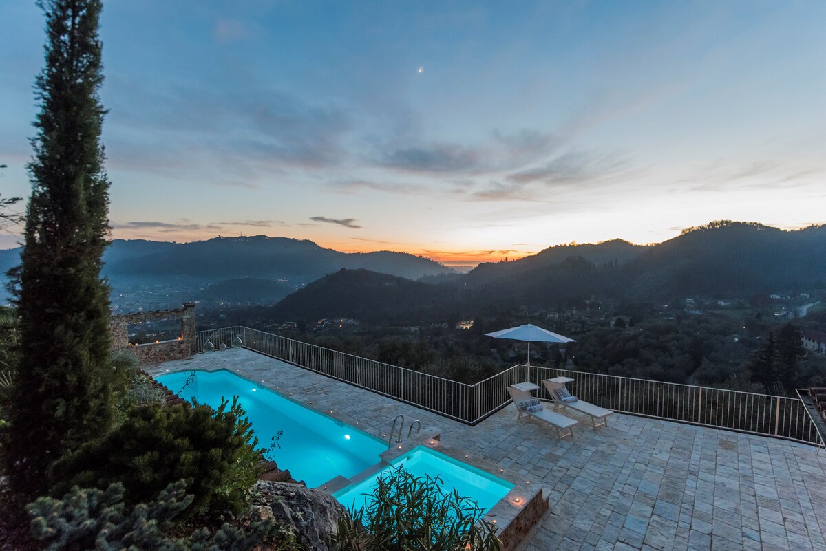 The image showcases a serene outdoor space featuring a swimming pool illuminated under the soft light of dusk. Surrounding mountains and valleys create a tranquil backdrop, while lounge chairs offer a comfortable spot for relaxation. A large umbrella provides shade, enhancing the inviting atmosphere.