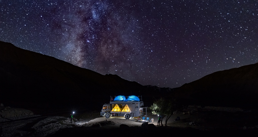A campervan is positioned under a clear night sky filled with stars, showcasing a view of the Milky Way. The van is illuminated softly, with blue tents set up on the roof. Silhouetted mountain peaks frame the scene, creating a serene outdoor atmosphere.