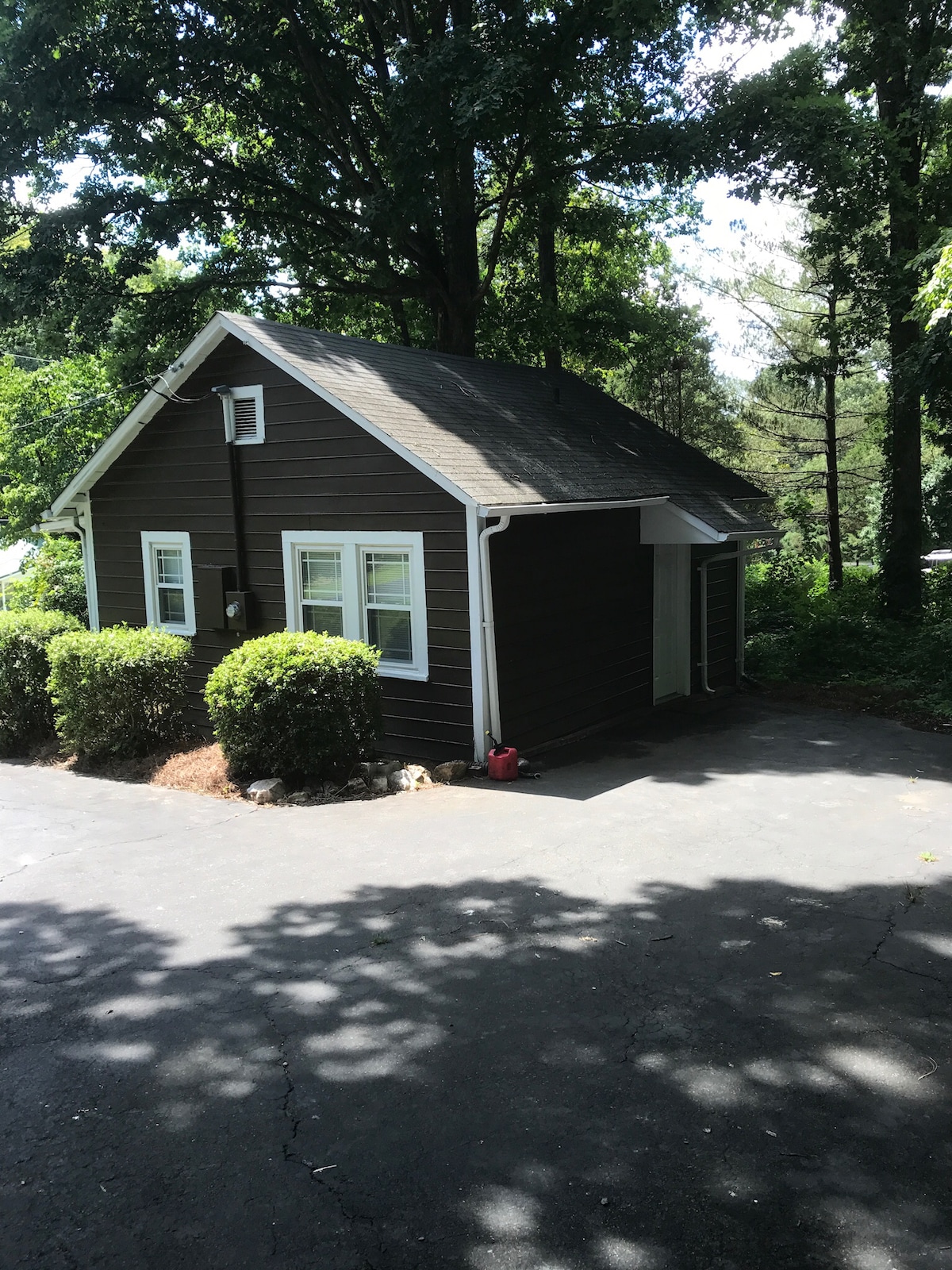 A cozy tiny house is nestled among lush greenery, featuring a simple brown exterior with white trim. Large windows allow natural light, while well-maintained bushes frame the entrance. The paved driveway leads to the private setting, surrounded by trees that provide a serene atmosphere.