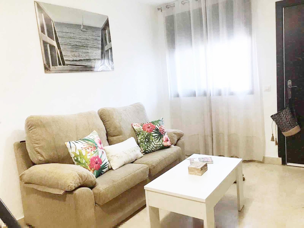 A cozy living area is presented with a light beige sofa adorned with floral throw pillows. A white coffee table is placed in front of the sofa, and a framed photo of a sailboat hangs on the wall. Natural light filters through sheer curtains at the window.
