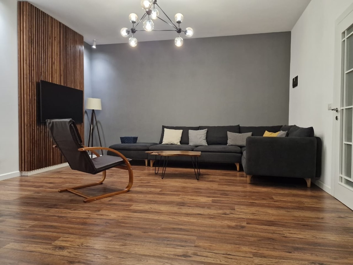 A modern living area features a dark sectional sofa with a mix of decorative pillows. A wooden coffee table sits in front, complemented by a contemporary rocking chair. The space is enhanced by a unique light fixture above and warm wooden flooring.