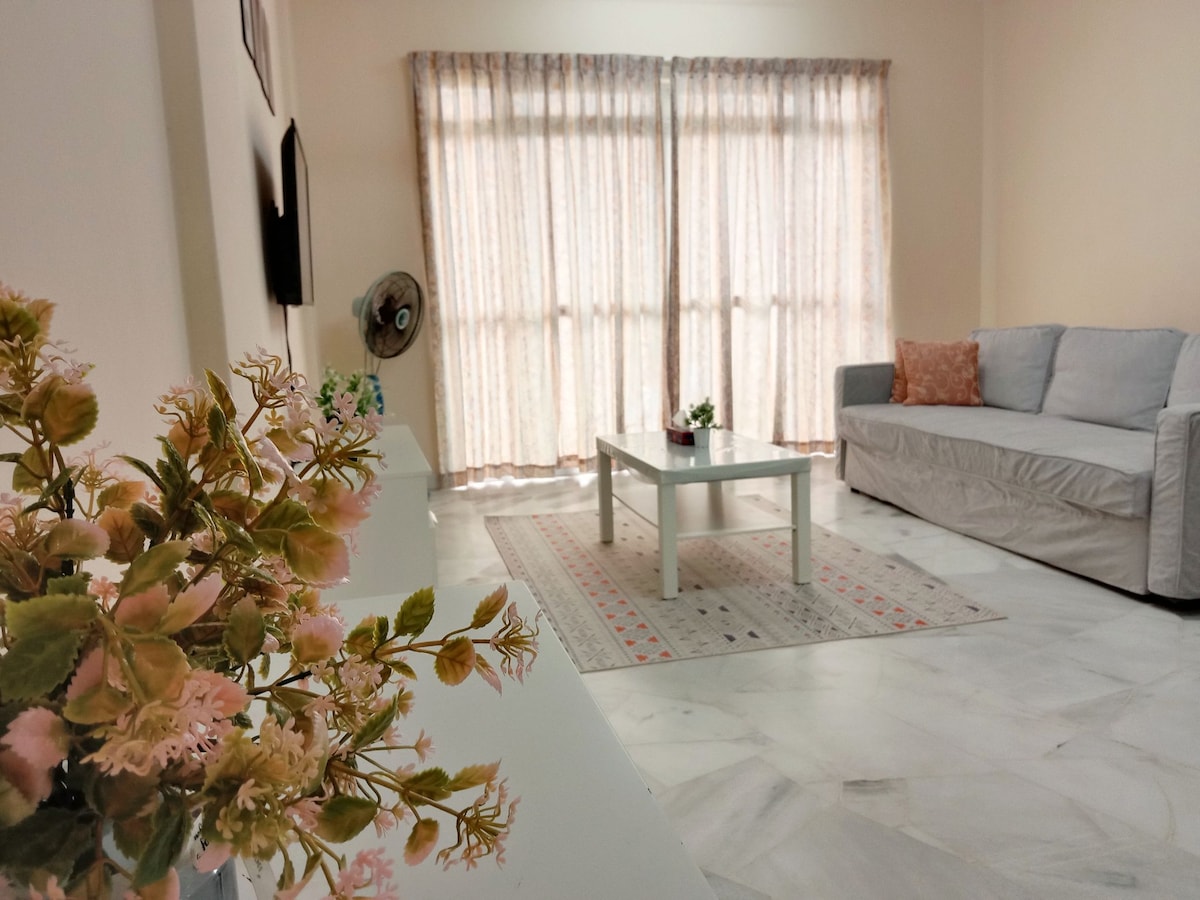 A bright living area features large windows with light curtains, allowing natural light to fill the space. A comfortable sofa is positioned near a low coffee table, surrounded by a decorative area rug. A potted flower arrangement adds a touch of greenery.