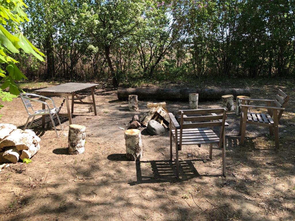 An outdoor fire pit area is surrounded by wooden stools and chairs, providing a cozy gathering space. A rustic wooden table is positioned nearby, while the ground is covered with a light layer of earth. Lush greenery encircles the area, adding a sense of privacy.