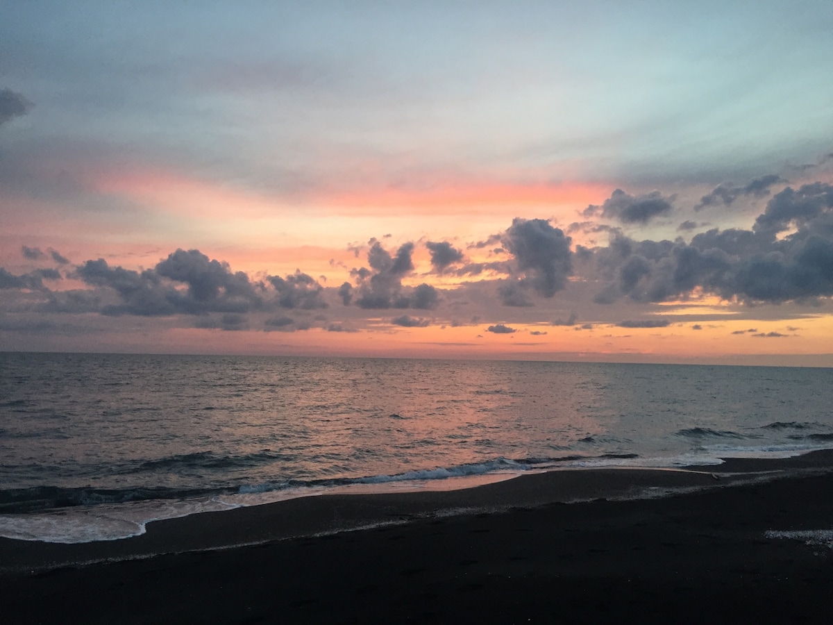 The image captures a serene beach scene at sunset, where soft pink and orange hues blend seamlessly in the sky above gentle waves. The dark shoreline contrasts with the colorful horizon, creating a tranquil atmosphere by the sea.