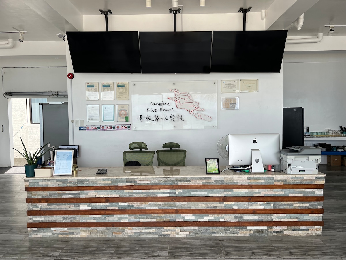 The reception area features a modern front desk constructed with a mix of stone and wooden accents. Behind the desk, two green chairs are positioned, and a large wall-mounted display houses documents and certificates. The space is well-lit and organized, contributing to a welcoming entry point.