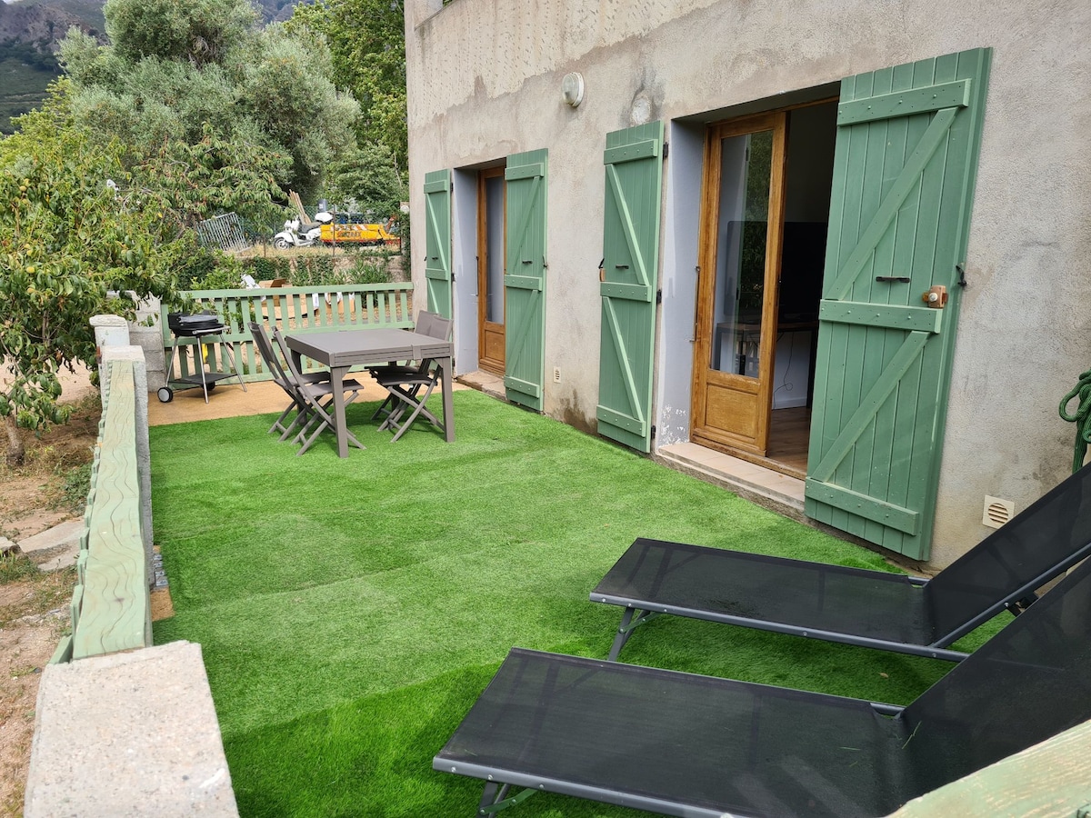A private terrace features green artificial grass with a dining table and chairs, complemented by two lounge chairs positioned for relaxation. Wooden shutters frame the doors, leading into the interior space, while surrounding greenery enhances the tranquil environment.