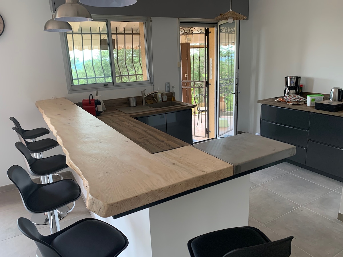 A modern kitchen features a wooden breakfast bar with sleek black stools arranged around it. Large windows provide natural light and offer views of the outdoor area. Coffee appliances are placed on the countertop, contributing to a functional and open space.