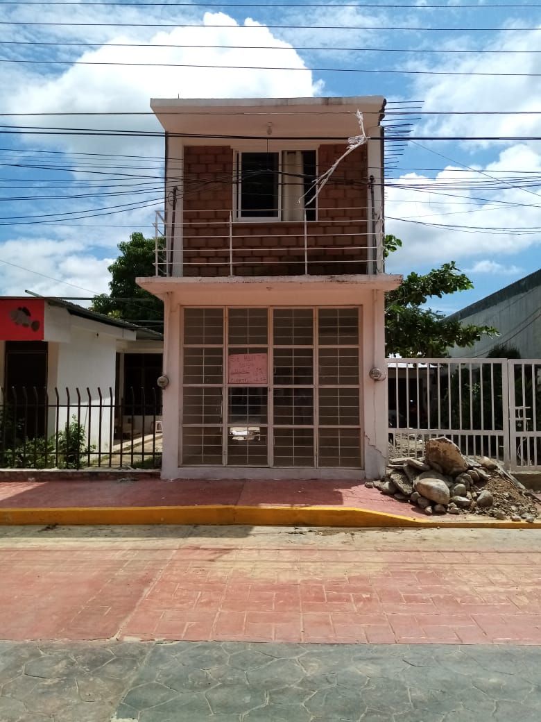 The exterior of a two-story building is presented, featuring a simple brick facade and large glass windows secured by metal grilles. A low fence frames the property, with a paved walkway leading to the entrance. The scene includes greenery in the background under a partly cloudy sky.