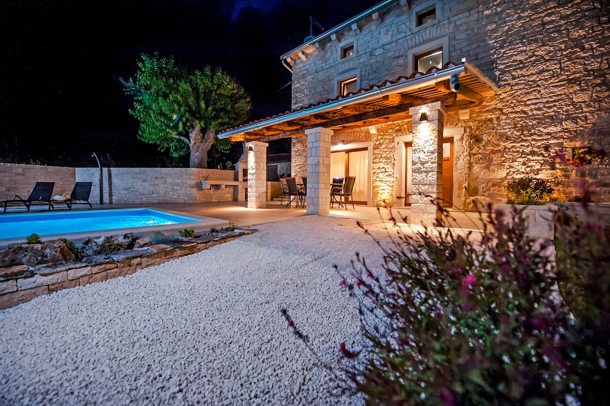 The exterior of a traditional stone house is illuminated at night, showcasing a swimming pool surrounded by a gravel area. A covered terrace with seating is visible, alongside landscaped greenery and flowers that enhance the outdoor space.