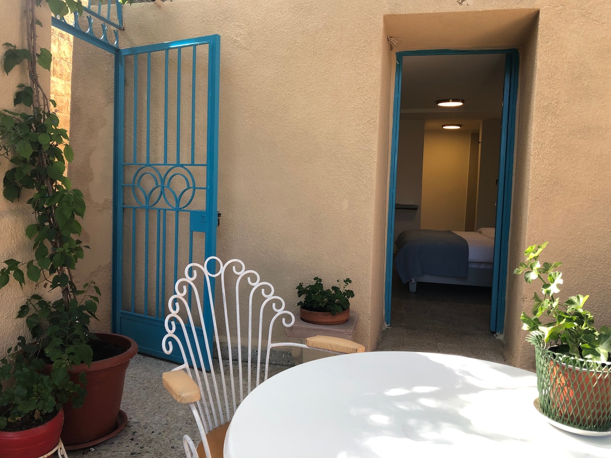 An outdoor terrace is highlighted by a circular white table and an ornate metal chair. Potted plants adorn the space, while a blue door leads into the minimalist interior of the apartment, inviting light and a sense of openness.