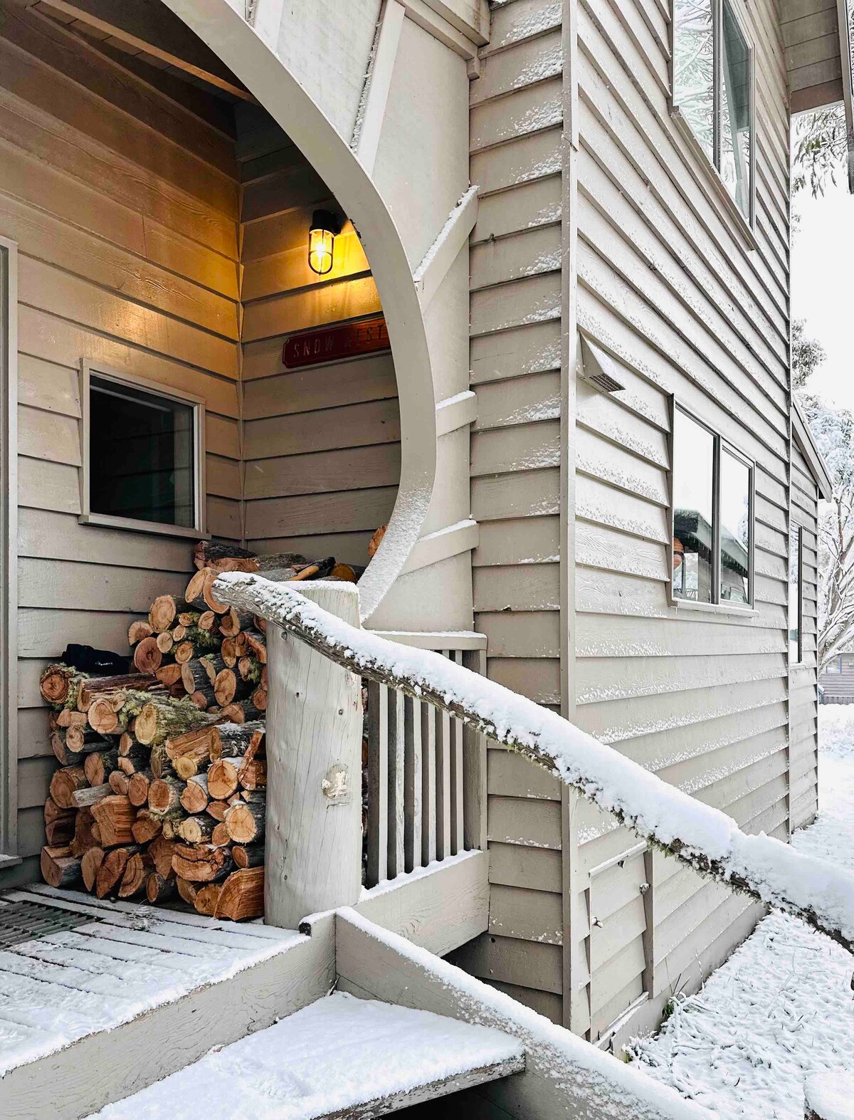 The chalet's entrance is showcased, featuring a cozy staircase leading to the front door. A neatly stacked collection of firewood is positioned beside the entrance, surrounded by a light dusting of fresh snow.