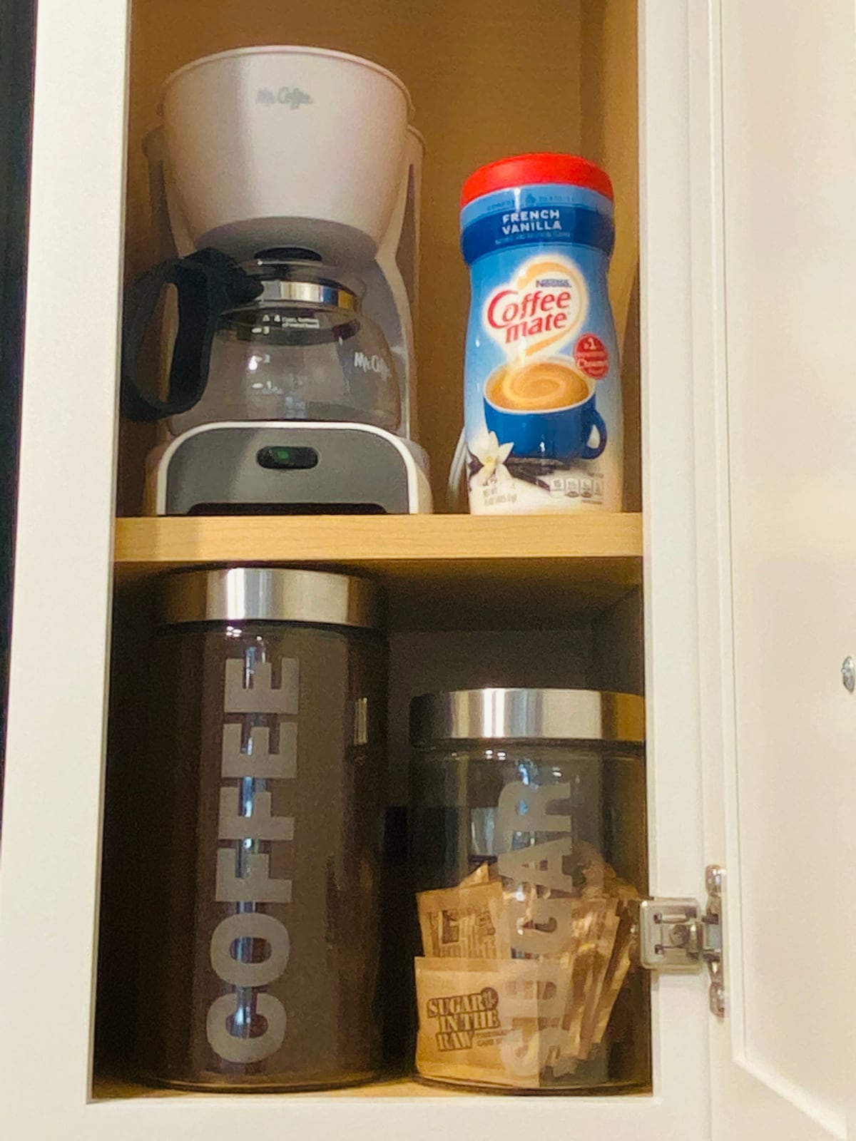 An organized cupboard displays a coffee maker beneath a container labeled 'COFFEE' and a glass jar for 'SUGAR.' Creamer is placed prominently beside the coffee maker, providing essentials for a morning beverage.