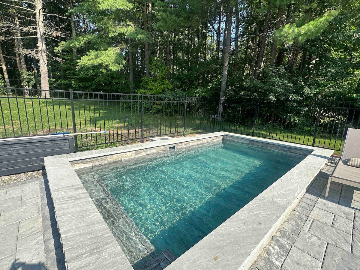 A private plunge pool with a sleek stone border is set within a fenced area. Clear, inviting water reflects surrounding greenery, creating a serene atmosphere. Adjacent lounge chairs provide comfortable seating for relaxation in a tranquil wooded setting.